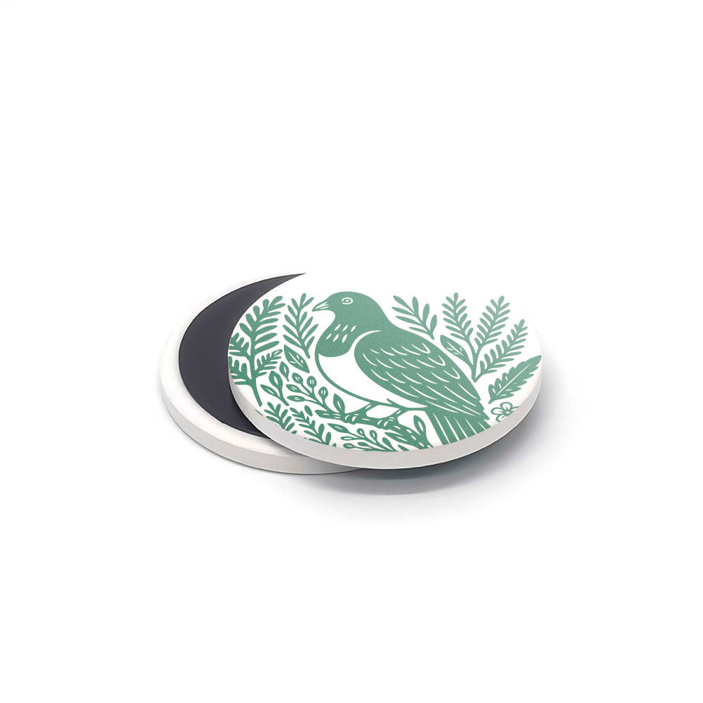A round, white ceramic magnet from the Woodcut Ceramic Magnet Set features a green bird and foliage design, overlapping a plain dark magnet—both perfect as New Zealand souvenirs and shown on a white surface.