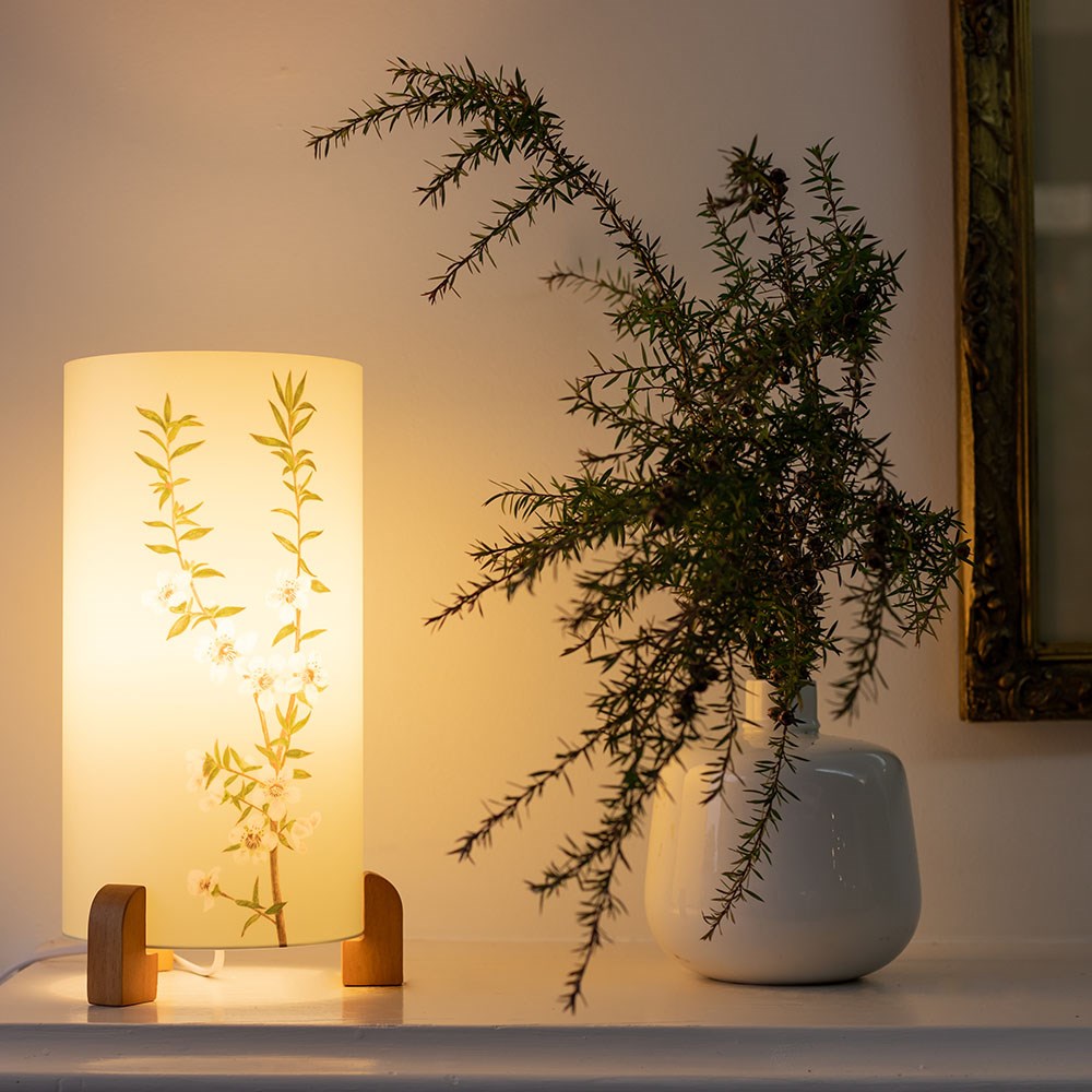 The Vintage Botanical Manuka Table Lamp sits beside a white vase with a leafy branch on a white surface against a pale wall, evoking vibrant scenes from stylish New Zealand interiors.