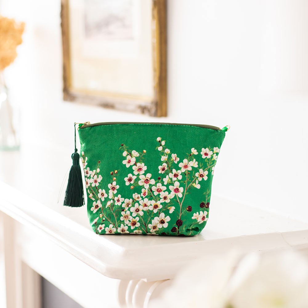 The Manuka Flower Velvet Cosmetic Bag, featuring a white and red Manuka flower pattern and a dark green tassel, rests on a white surface in a bright room with a framed picture in the background.