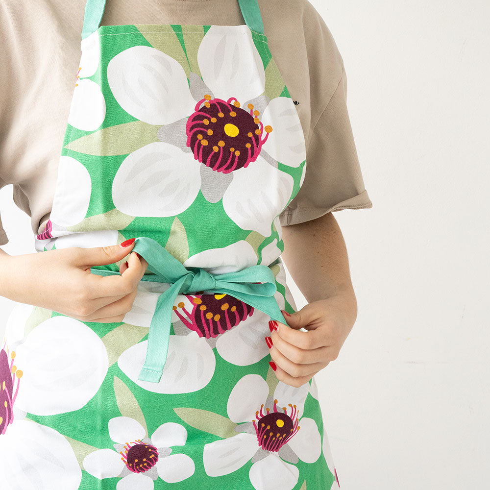 A person ties the waist strings of a green Bright Botanical Manuka Apron made from 100% cotton, with a white and purple flower pattern. Only their torso and hands are visible against a plain white background; they wear a beige shirt.