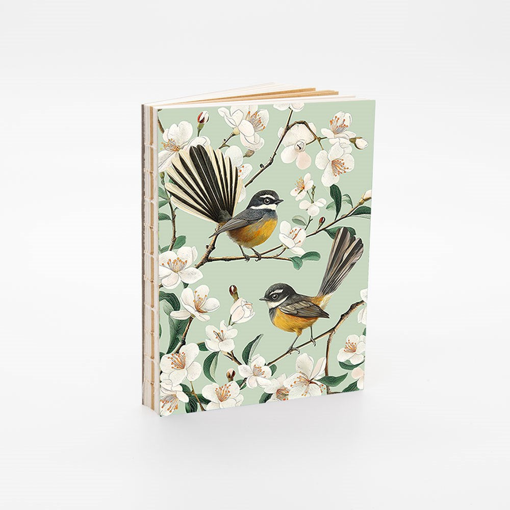 The Chatty Fantails Notebook is a hardcover art notebook with a pale green cover featuring two birds on blossoming branches with white flowers, shown upright against a white background. It includes unlined pages for creative freedom.