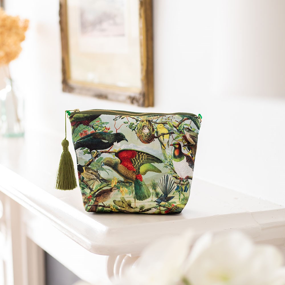 The NZ Native Birds Velvet Cosmetic Bag, featuring a colorful print of New Zealand birds and foliage and a green tassel zipper, sits on a white mantel with charming accents like a blurred framed picture and dried flowers in the background.