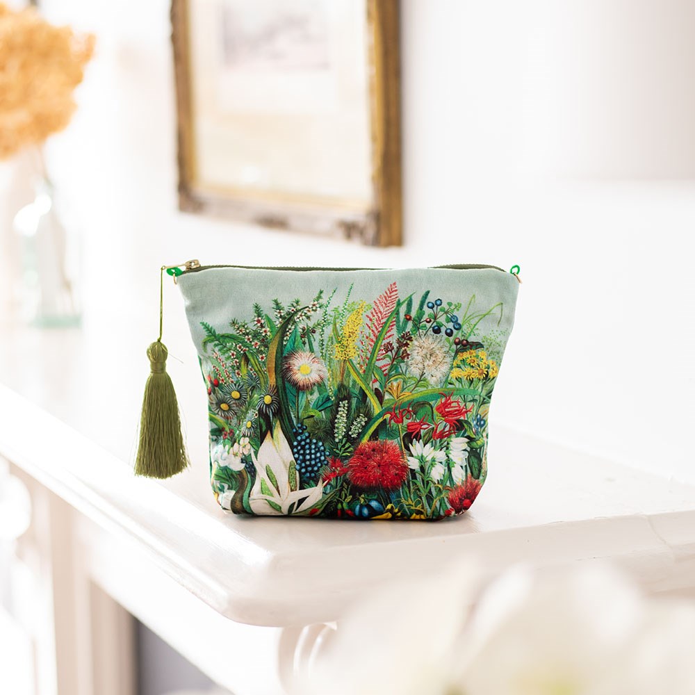 The NZ Native Flowers Velvet Cosmetic Bag, featuring a colorful botanical design by New Zealand artists and a green tassel on the zipper, sits on a white mantel—an ideal gift with blurred decor and framed art in the background.