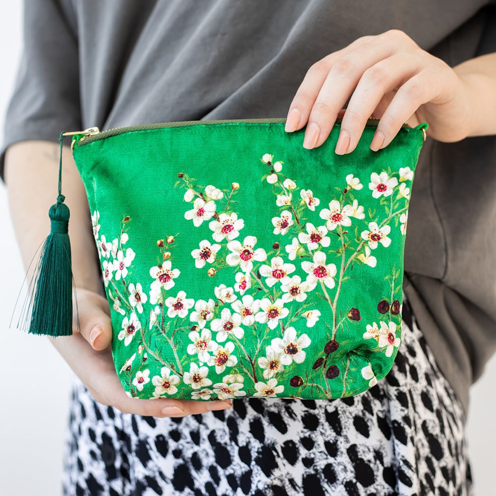 A person holds the Manuka Flower Velvet Cosmetic Bag, featuring white and pink floral embroidery and a green tassel, paired with a grey top and black-and-white patterned skirt, capturing the charm of New Zealand.