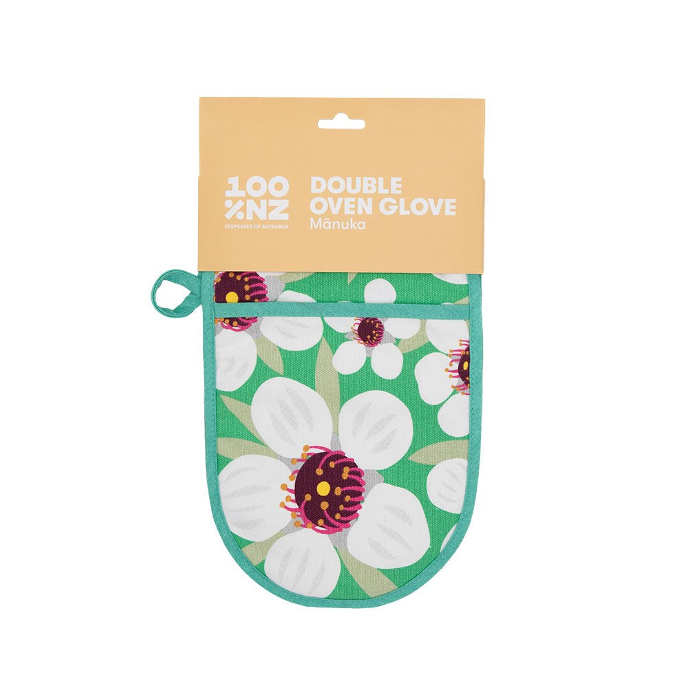 The Bright Botanical Manuka Double Oven Glove, made from 100% cotton, features a green background with white and purple floral patterns. Shown in its packaging, it’s labeled as 100% NZ Double Oven Glove Manuka.