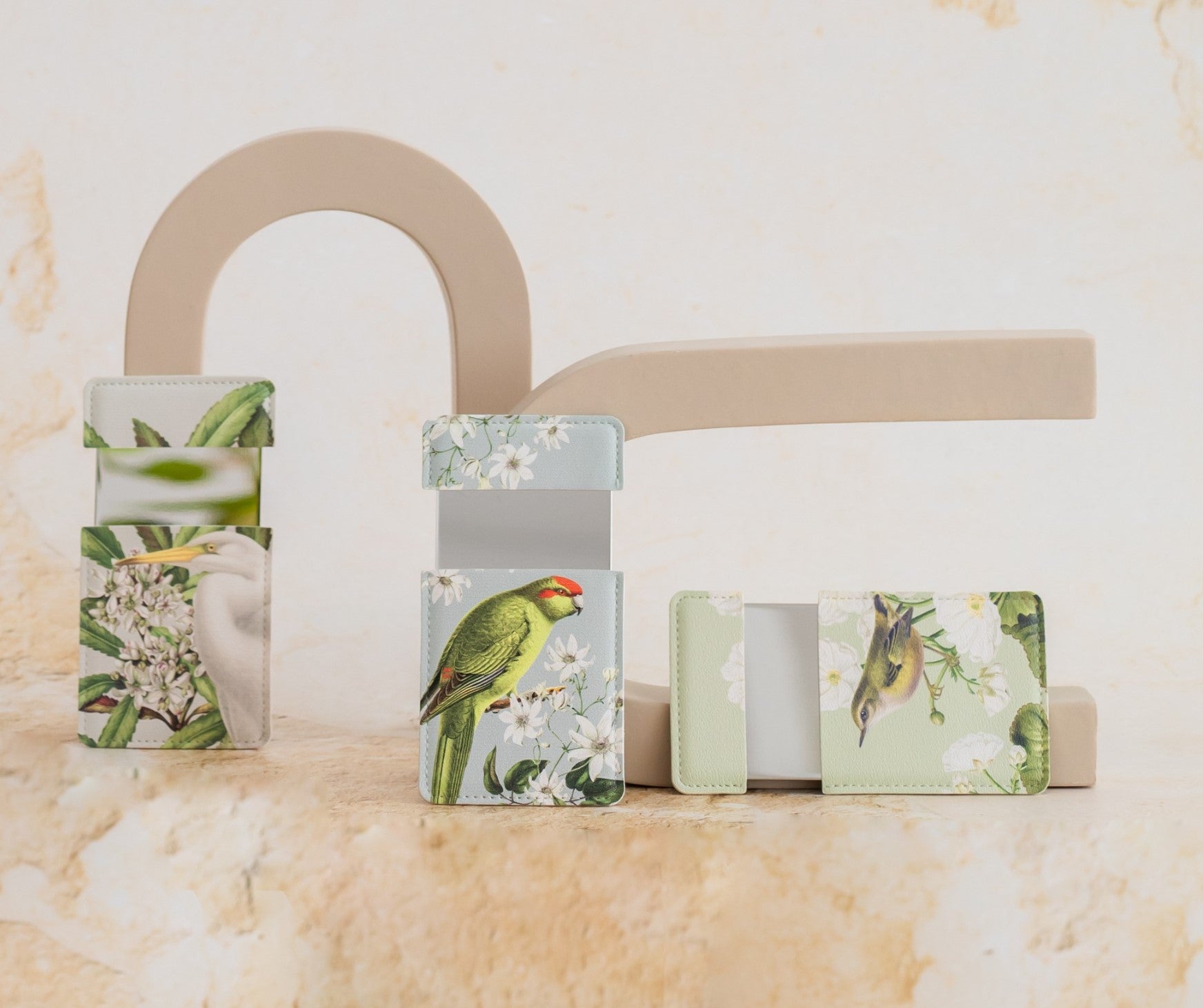 A pastel-colored vegan leather Birds & Botanics Heron Pocket Mirror is displayed on beige geometric props against a light, textured background, shown open to reveal its interior.