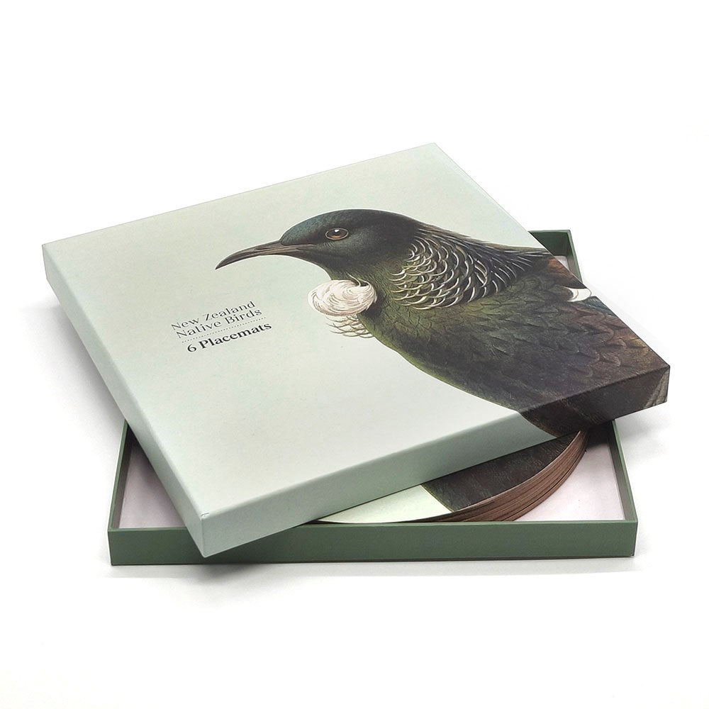 A light green box labeled Hushed Birds Box of 6 placemats features a realistic Tūī bird illustration on the lid. The partially open lid reveals six round placemats adorned with beautiful native bird designs inside.