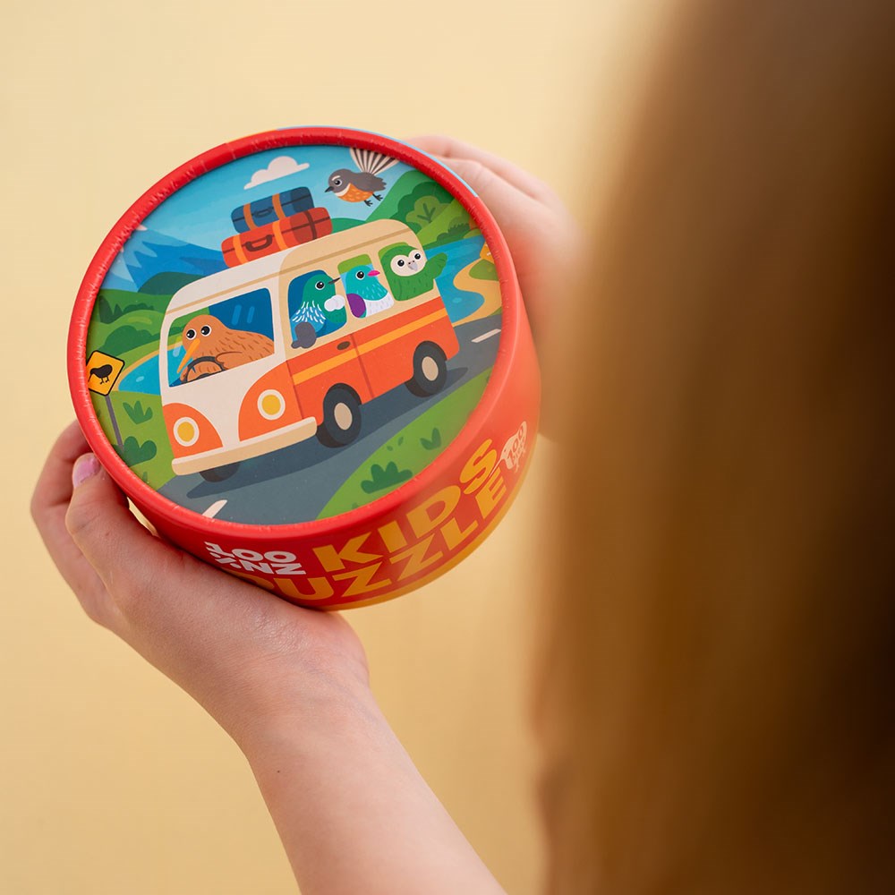 A child holds a round, red container with a lively lid featuring animals like a walrus, birds, and sloth in a van. The Kiwi Road Trip Kids Puzzle promises New Zealand-themed fun for children.