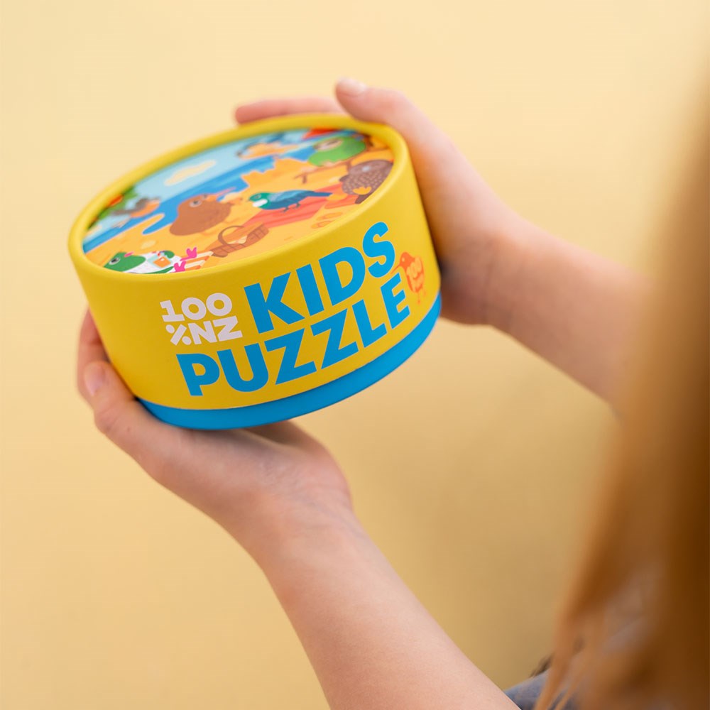 A child holds a round, yellow and blue box labeled Beach Party Kids Puzzle featuring native birds of New Zealand on the lid, against a beige background.