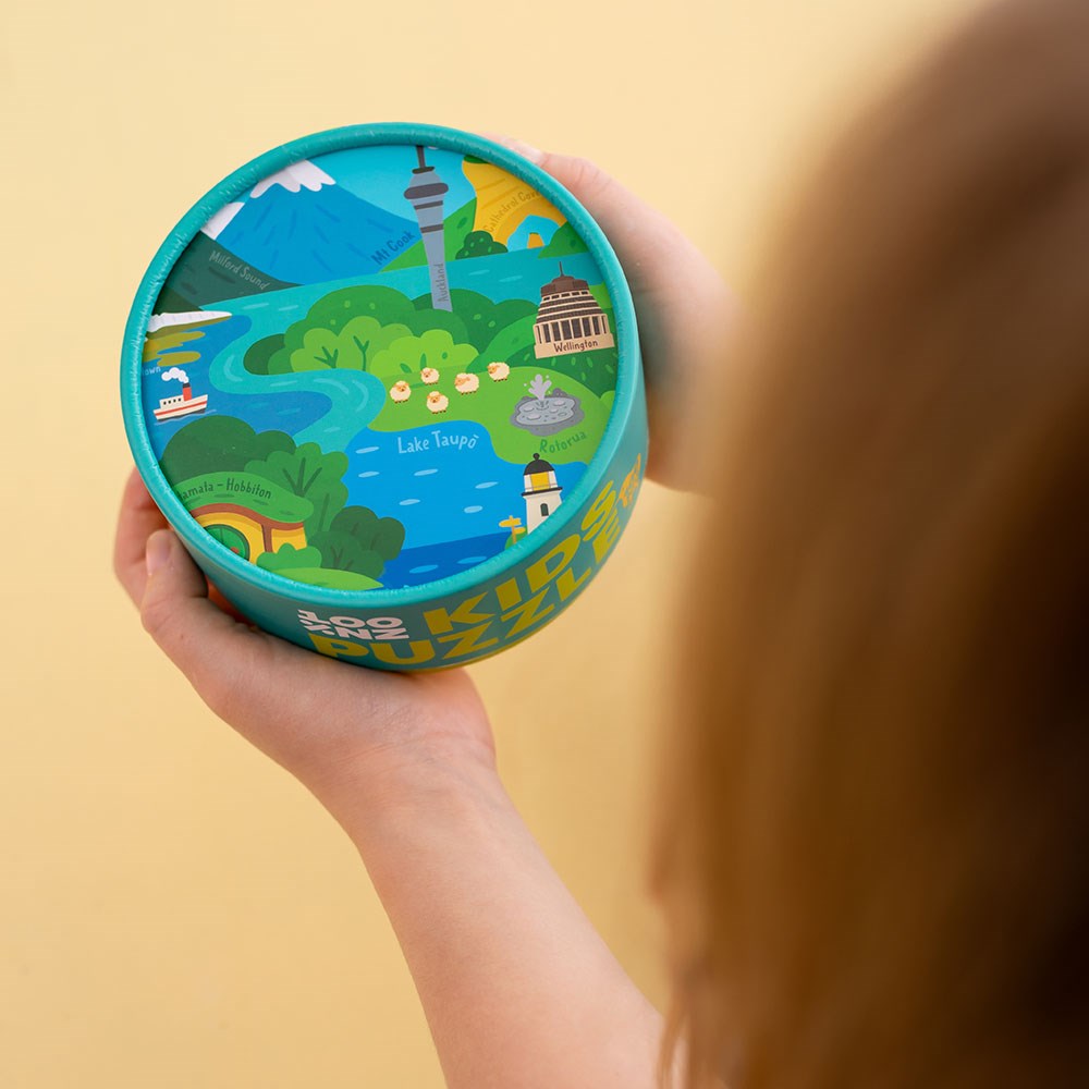 A child holds the NZ Highlights Kids Puzzle—a round blue container with a map of New Zealand landmarks like Lake Taupō, Rotorua, and Auckland—perfect for inspiring curiosity through fun puzzle play.
