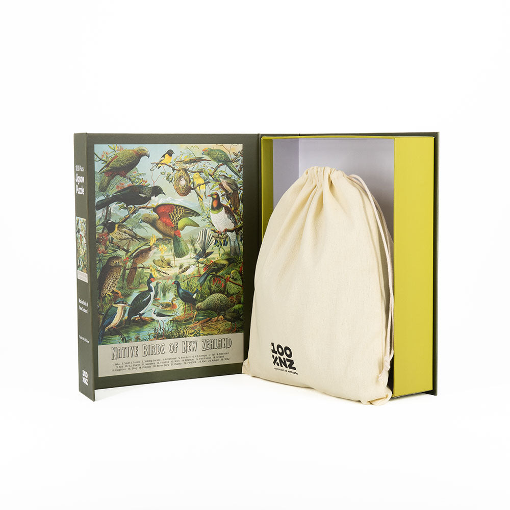 An open box reveals the NZ Native Bird Jigsaw Puzzle (1000 pieces), an illustrated book cover featuring native birds, and a cream-colored drawstring bag.