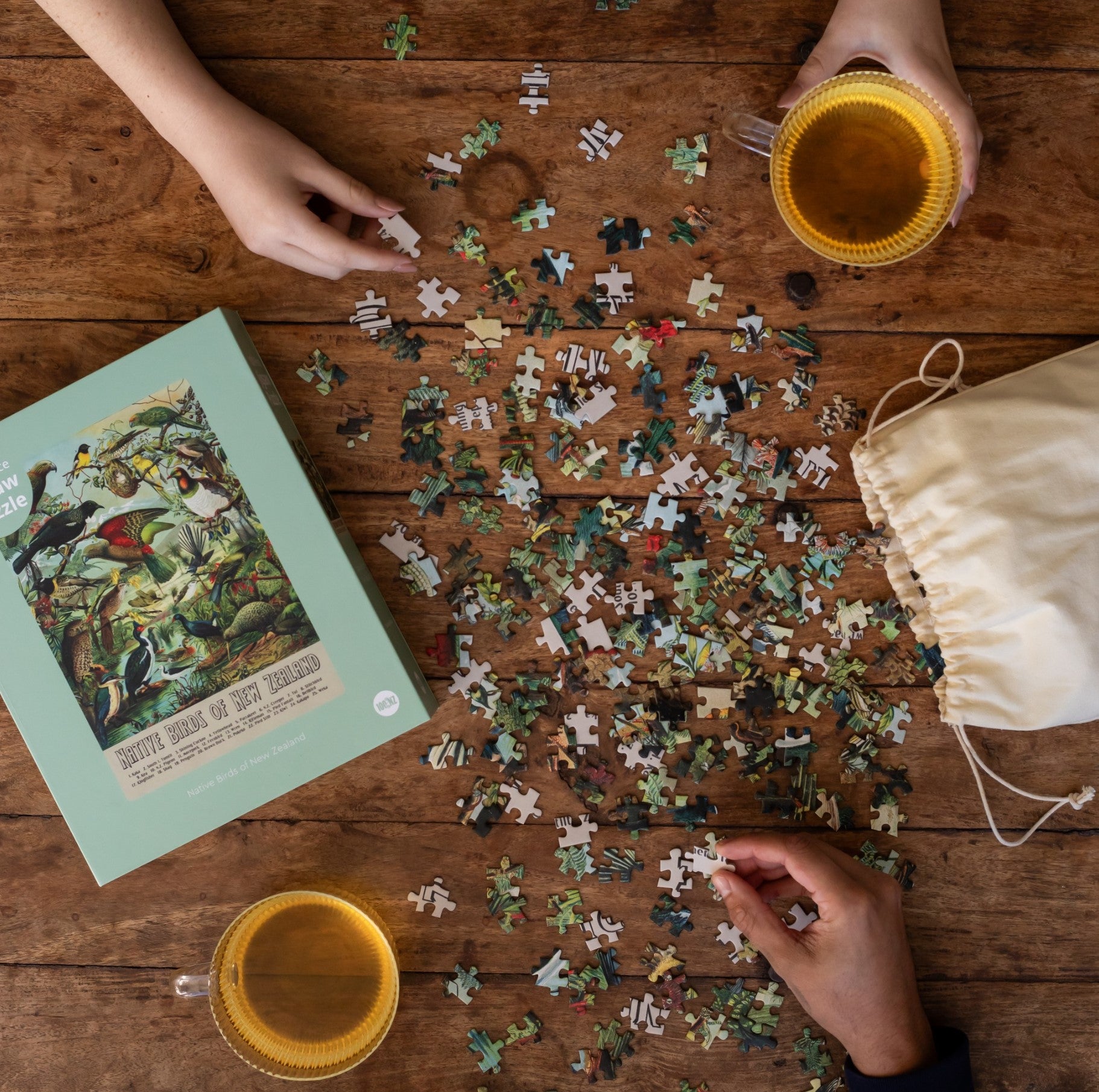 Two people assemble the NZ Native Bird Jigsaw Puzzle Box on a wooden table, surrounded by birdlife art, two cups of tea, a drawstring bag, and the puzzle box for a cozy atmosphere.