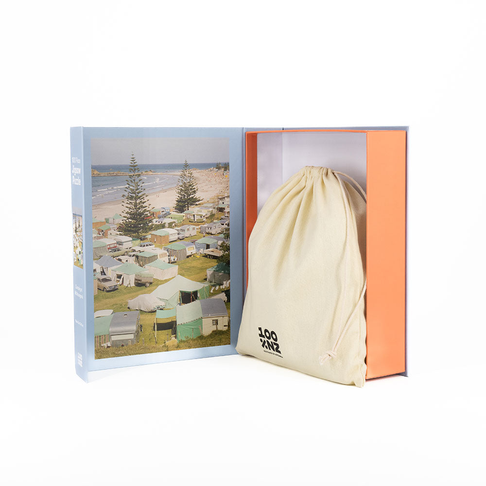 An open box displays the Camping at Mt Maunganui Jigsaw Puzzle, featuring a hardcover-style lid with a coastal scene and a cloth drawstring bag in the right compartment printed with black text. The background is white.
