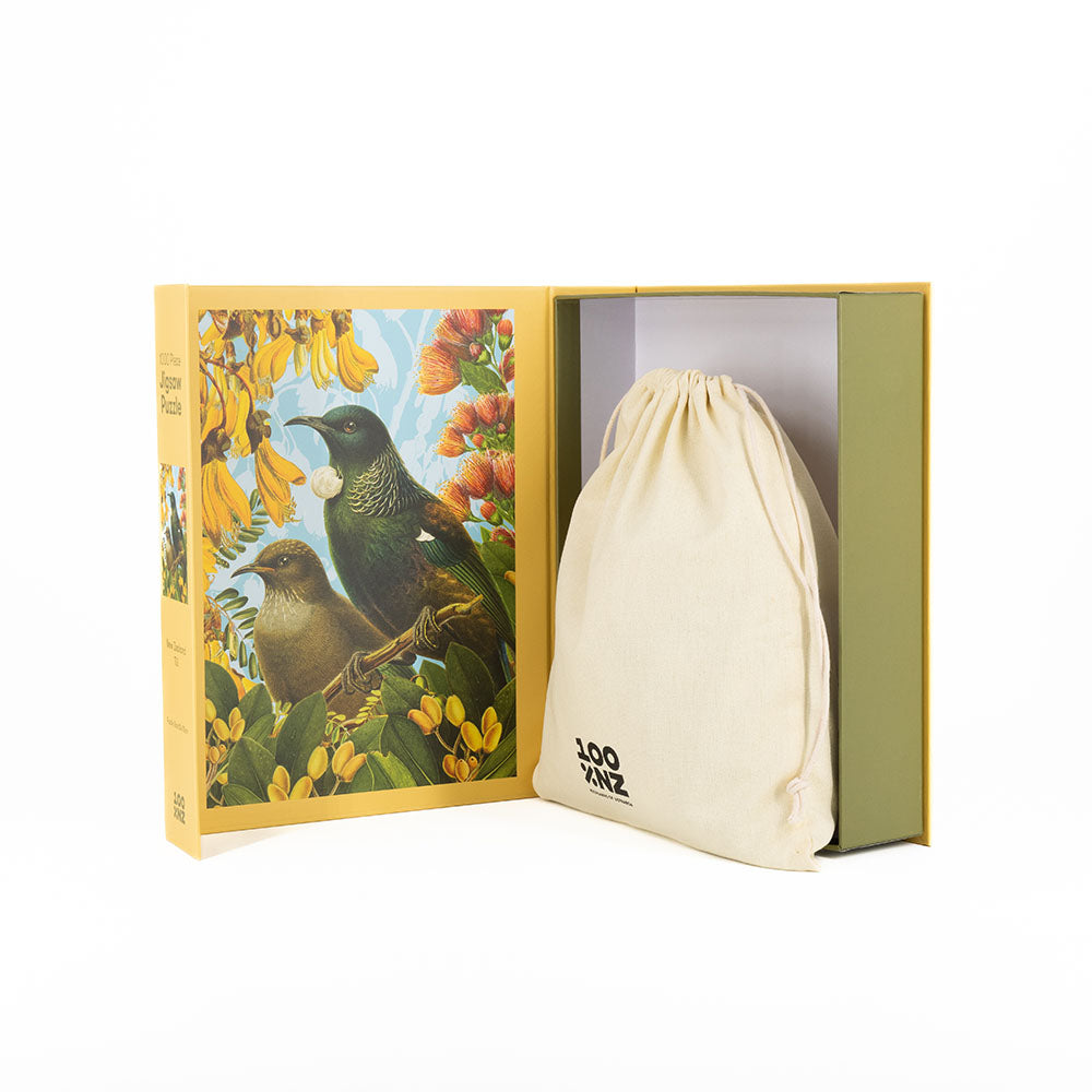 A yellow Botanical Tui Jigsaw Puzzle box with two New Zealand birds and yellow flowers is open, revealing a beige drawstring bag inside. The bag features black text that reads “100 XNZ.”.