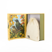 A yellow Botanical Tui Jigsaw Puzzle box with two New Zealand birds and yellow flowers is open, revealing a beige drawstring bag inside. The bag features black text that reads “100 XNZ.”.