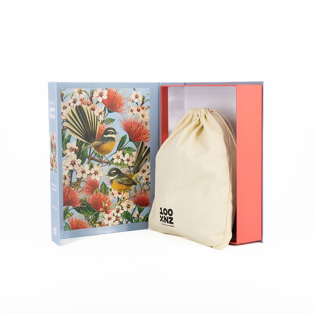 An open Native Flowers and Chatty Fantails Jigsaw Puzzle Box displays a cream drawstring bag with “LOO NZOO” partially visible, ideal for storing your 1000-piece puzzle—set against a white background.