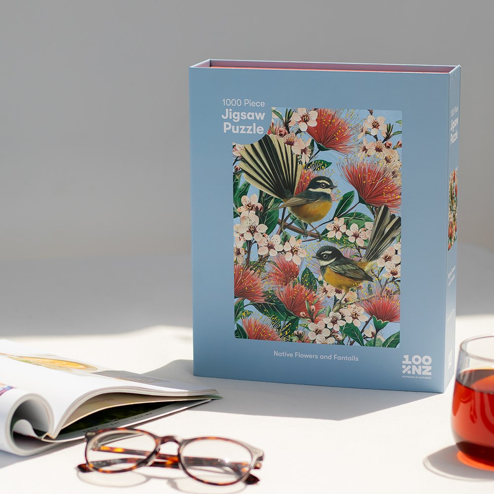 The Native Flowers and Chatty Fantails Jigsaw Puzzle Box (1000 pieces) rests on a white surface beside an open magazine, eyeglasses, and a glass of tea—the perfect setup for enjoying New Zealand’s native flowers and birds.