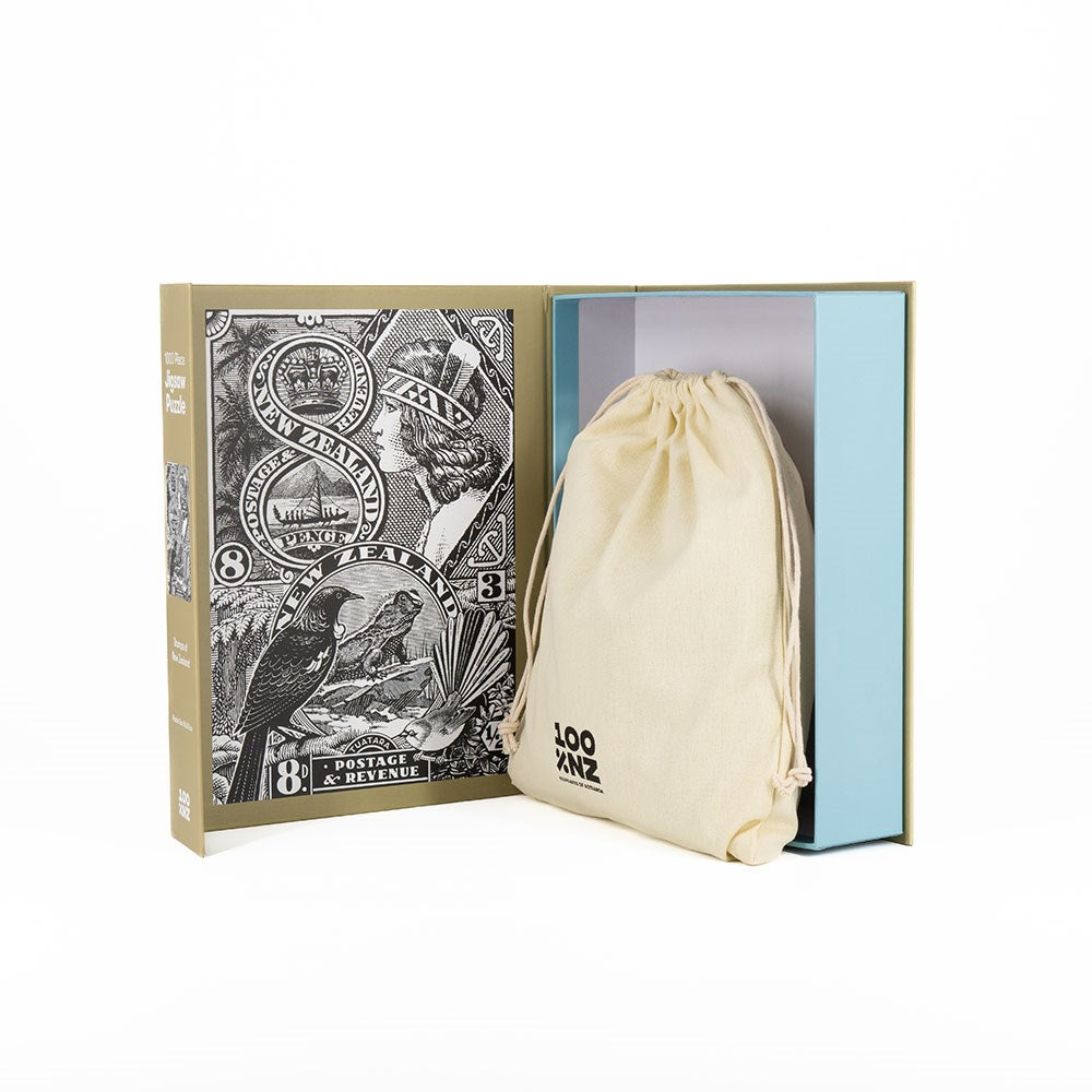 The Stamps of New Zealand Jigsaw Puzzle Box opens to show a cloth drawstring bag inside. The box’s left side displays detailed black and white vintage art of New Zealand bird stamps, adding an elegant collectible feel.