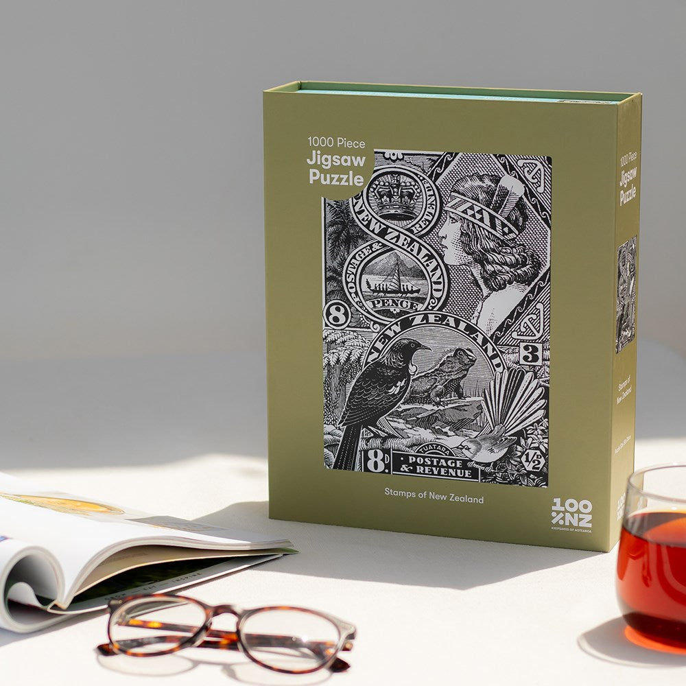 The Stamps of New Zealand Jigsaw Puzzle Box, a 1000-piece black and white vintage-inspired puzzle featuring New Zealand stamps, rests on a beige surface beside a book, eyeglasses, and a glass of tea.