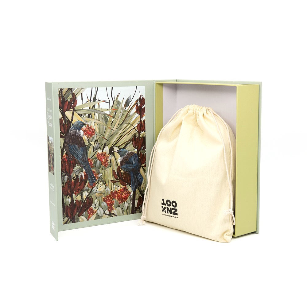The Kirsty Nixon - Double Bangers Jigsaw Puzzle comes in a light green and yellow box, with illustrated tui's and plants inside the cover and a LOOT-logo drawstring bag—an ideal keepsake for any New Zealand puzzle fan.