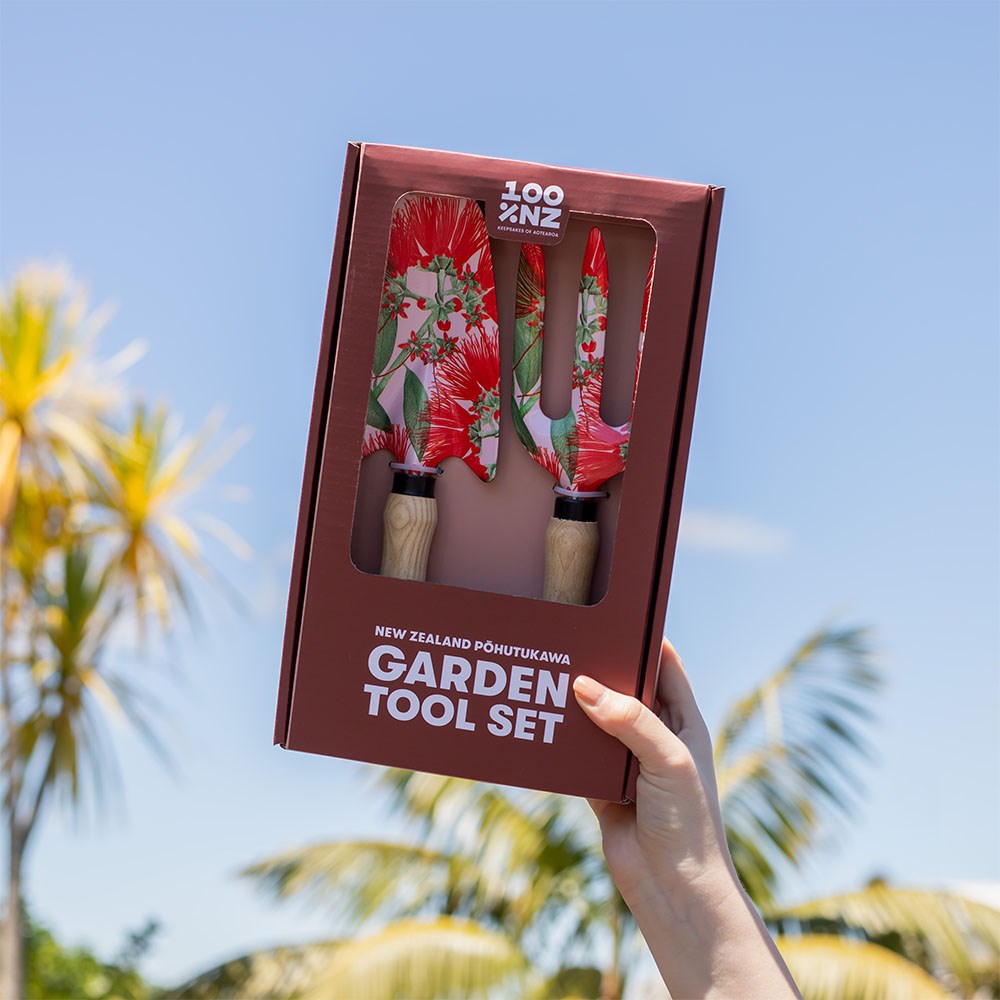 A hand holds up a boxed NZ Pohutukawa Garden Tool Set with floral-patterned handles, framed by palm trees and a blue sky.