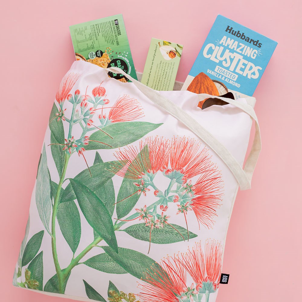 The Vintage Botanical Pohutukawa Shopping Bag, featuring a floral and green leaf design, holds boxes of food—including one labeled “Amazing Clusters”—and is displayed on a pale pink background.