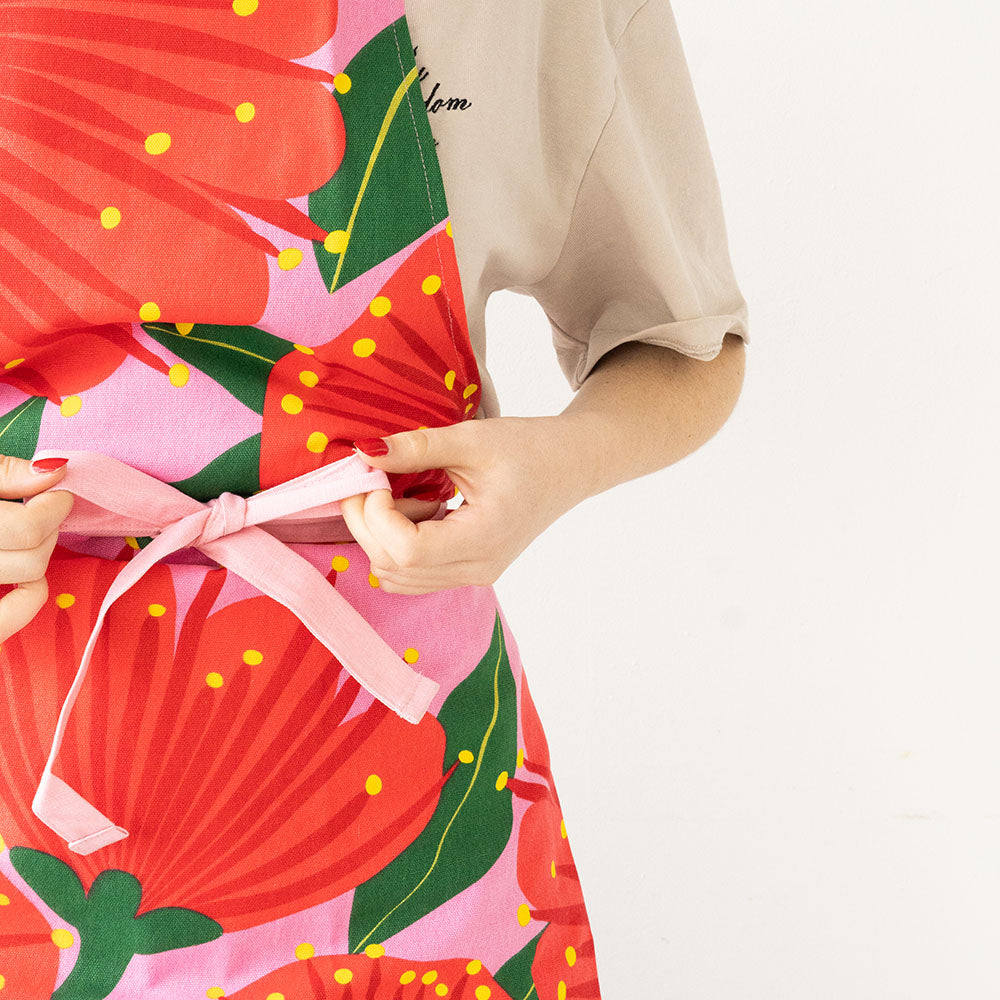 A person in a beige short-sleeve shirt ties the pink waistband of the Bright Botanical Pink Pohutukawa Apron (100% cotton) around their waist against a plain white background.