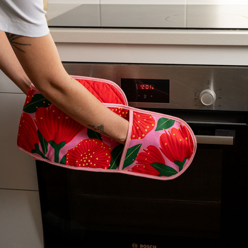 Wearing the Bright Botanical Pink Pohutukawa Double Oven Glove, a person opens a stainless steel oven showing 12:01. Only their arm is visible.