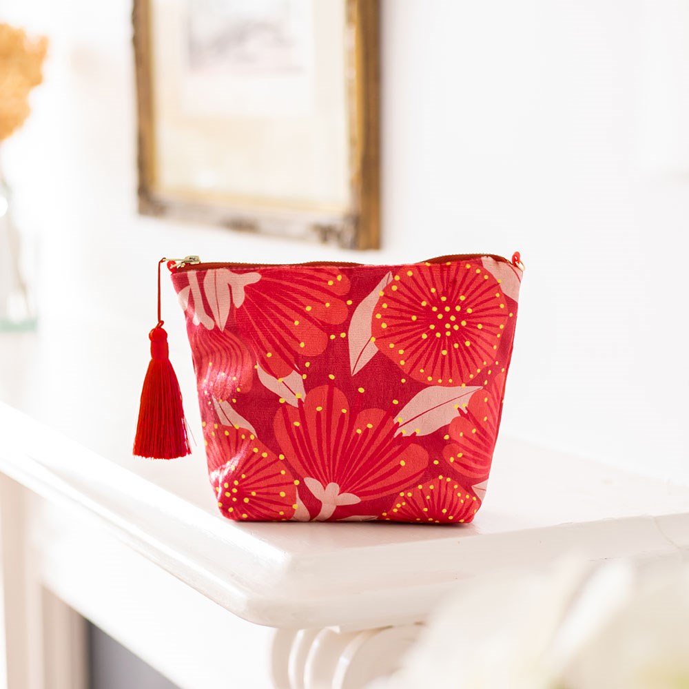The Bright Botanical Red Pohutukawa Velvet Cosmetic Bag, featuring a gold and light pink floral pattern and a red tassel, rests on a white mantel with a framed picture and blurred decor in the background.