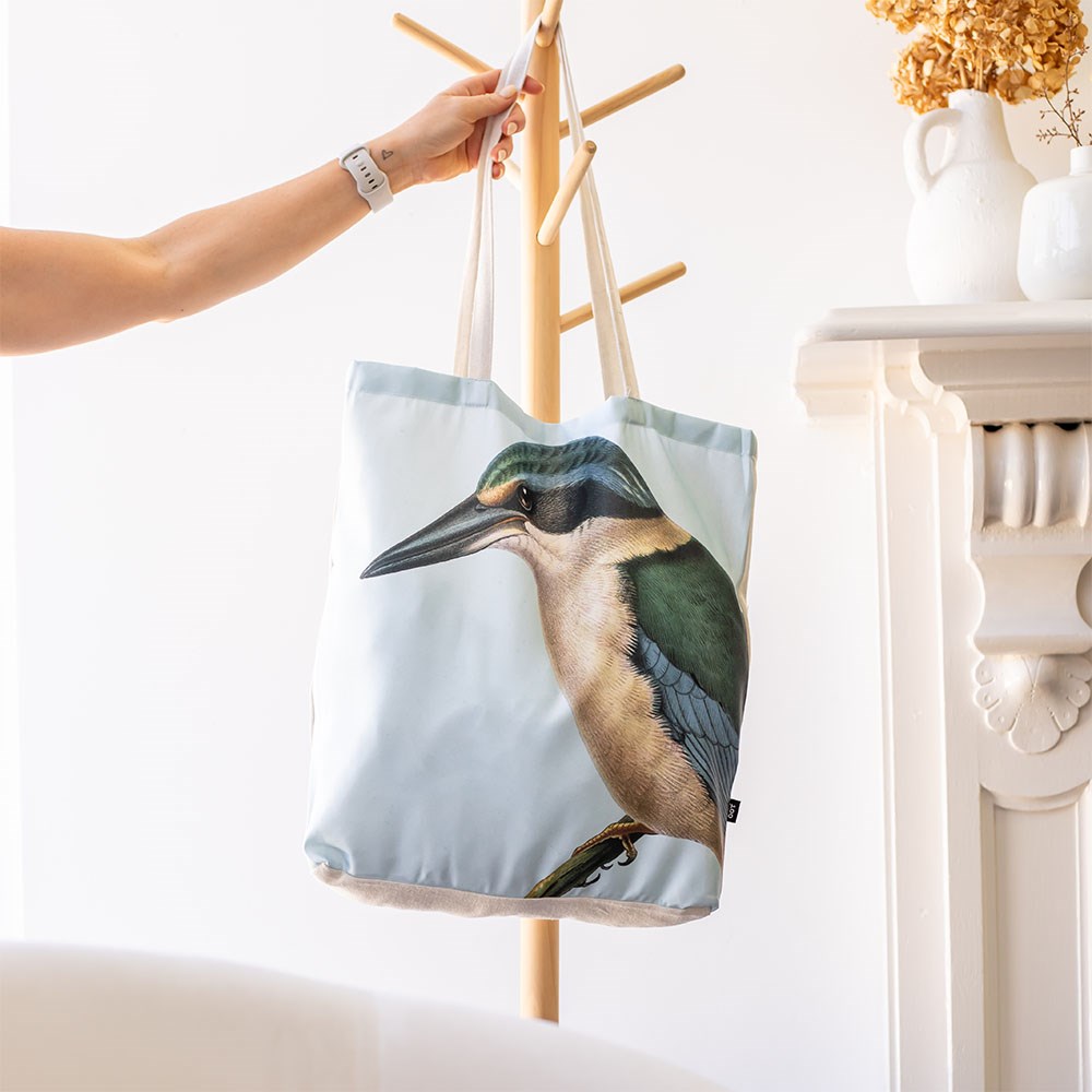 A person hangs the affordable Hushed Kingfisher Shopping Bag, featuring an illustrated kingfisher bird design, on a wooden coat rack beside a white fireplace with a vase of dried flowers.
