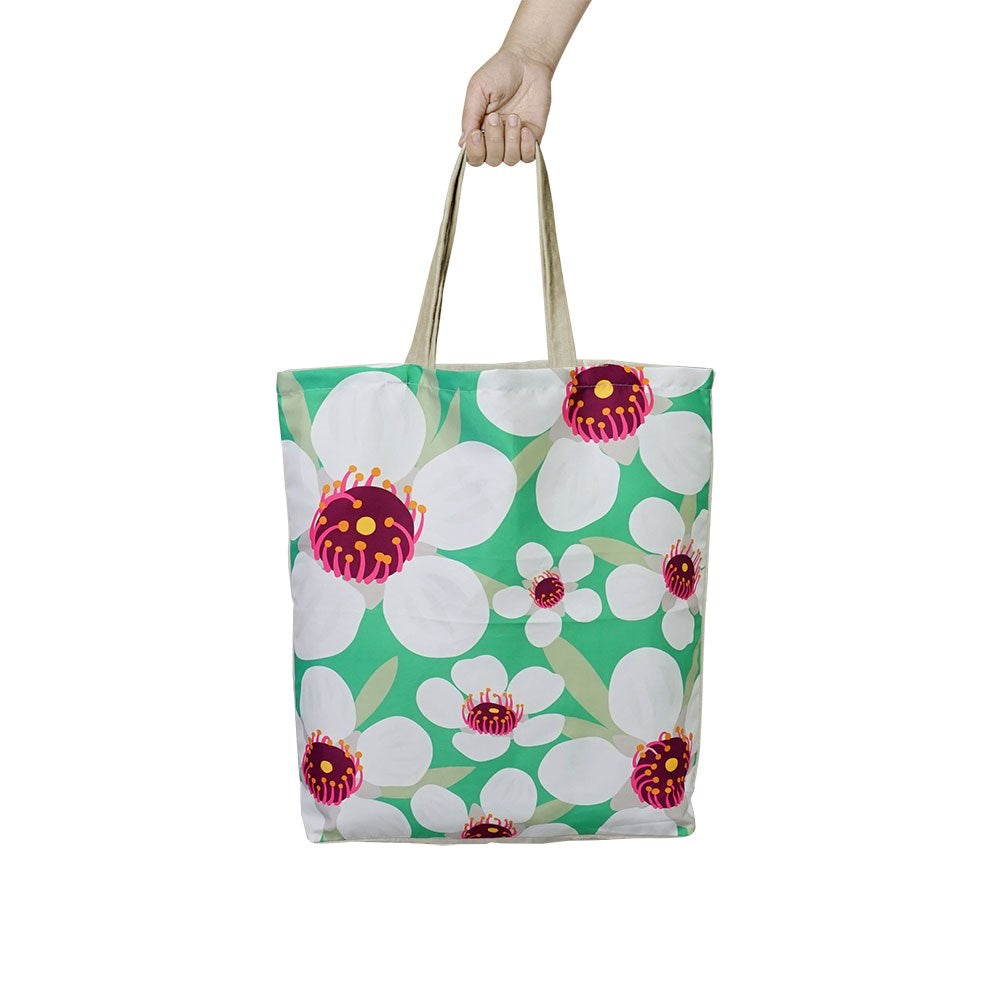 A hand holds the affordable Bright Manuka Shopping Bag, a reusable tote with beige handles and a green background decorated with large white flowers featuring pink and yellow centers.