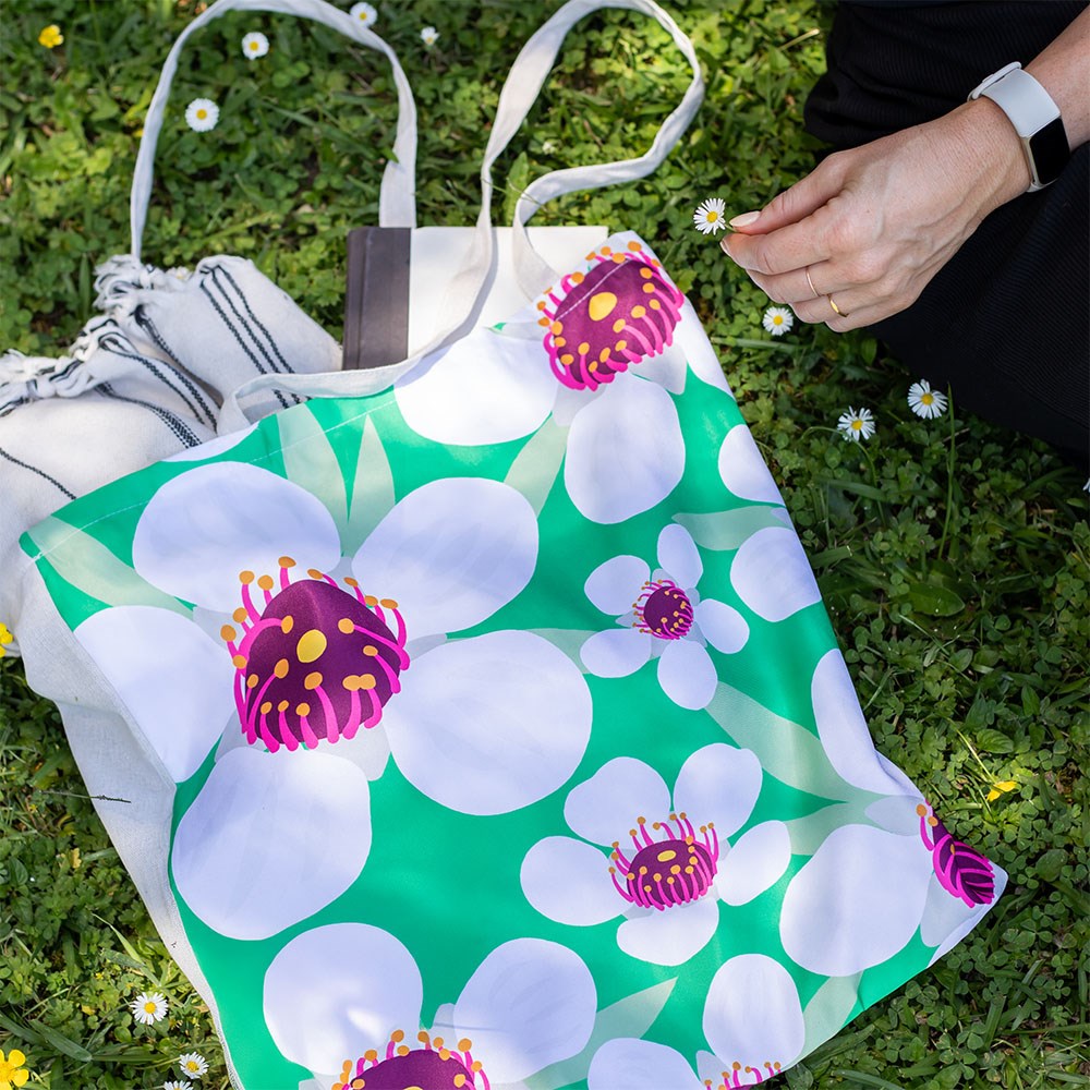 A person sits on grass holding a daisy, with the Bright Manuka Shopping Bag—a reusable tote featuring large white flowers with pink and yellow centers on green—a striped towel, and a book nearby.