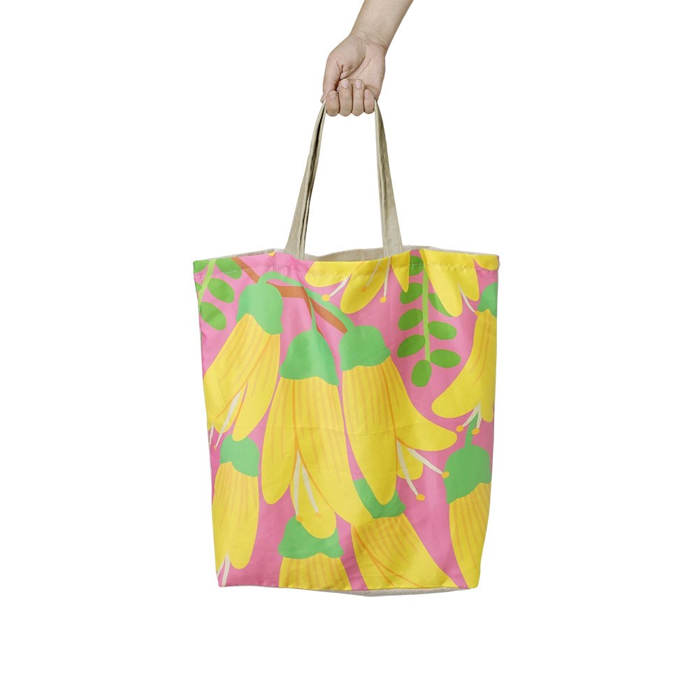 A hand holds the Bright Kowhai Shopping Bag, a lightweight reusable tote with beige handles. It features a vibrant pink background adorned with large yellow bananas and green leaves.