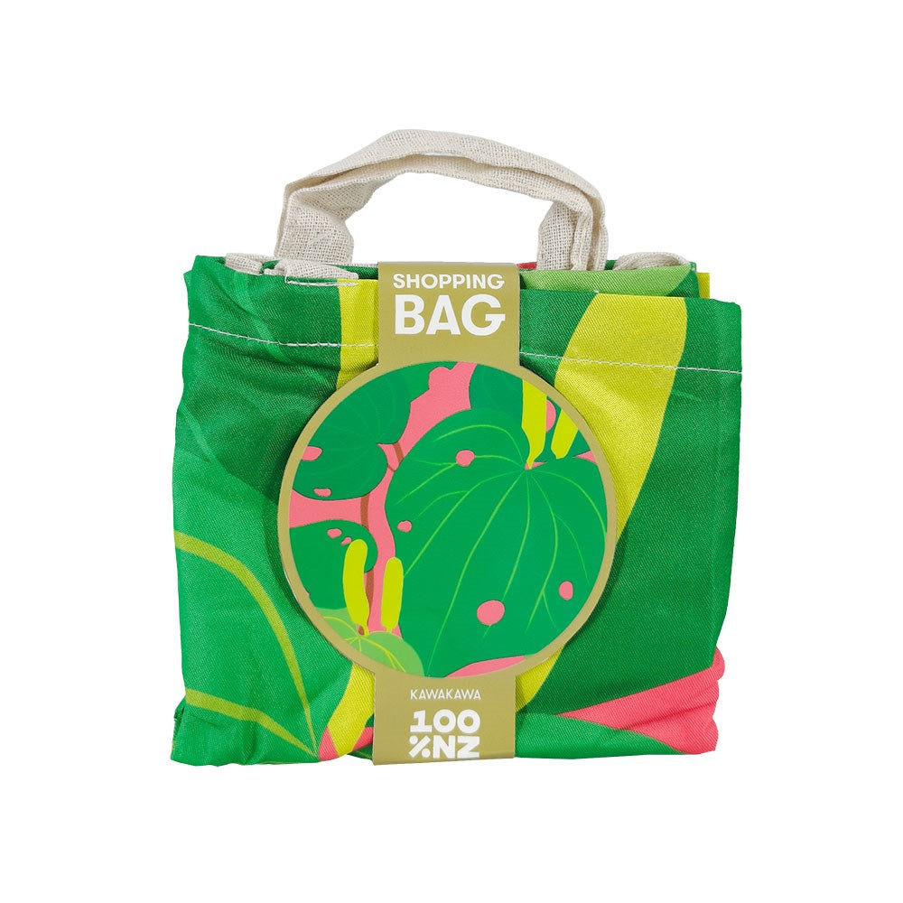 The Bright Kawakawa Shopping Bag is a lightweight, foldable, reusable bag with a green, yellow, and pink botanical pattern. It comes with short off-white handles and a round tag labeled Shopping Bag Kawakawa.