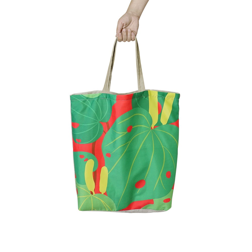 A hand holds the Bright Kawakawa Shopping Bag, a lightweight tote with beige handles and a bold design of large green leaves and yellow accents on a red background.