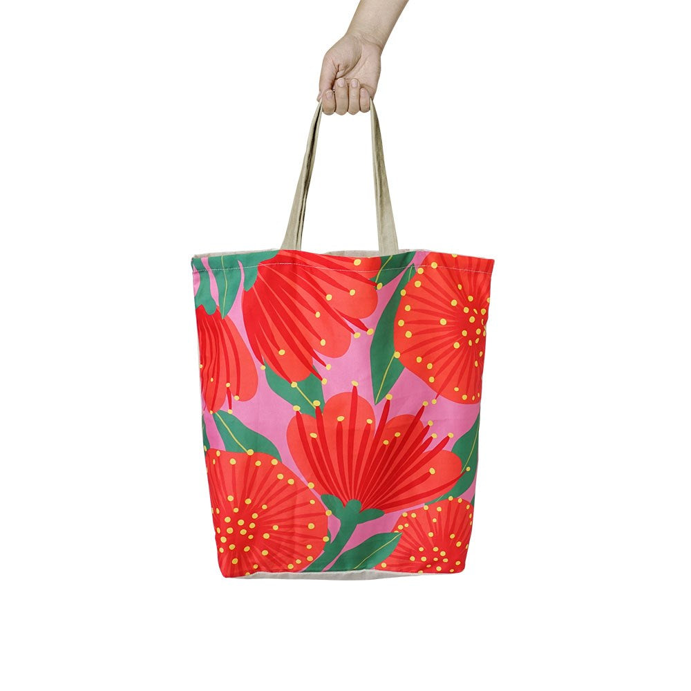 A hand holds the Bright Pink Pohutukawa Shopping Bag, an affordable, reusable tote with beige handles and a vibrant design of large red flowers and green leaves on a pink background—perfect for stylish shopping trips.