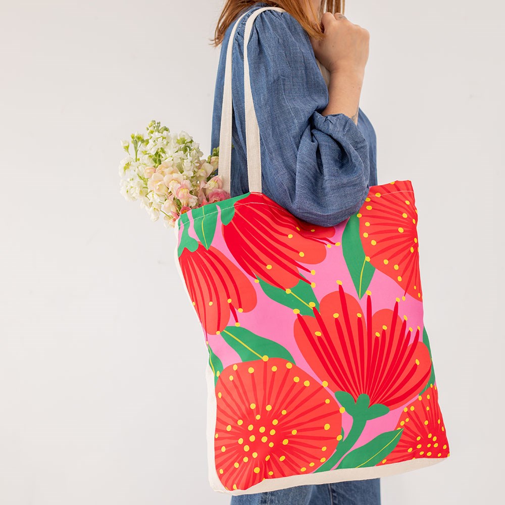 A person with long brown hair, wearing a blue long-sleeve top, carries an affordable Bright Pink Pohutukawa Shopping Bag decorated with red and yellow flowers on pink; white flowers peek out from inside the reusable tote.