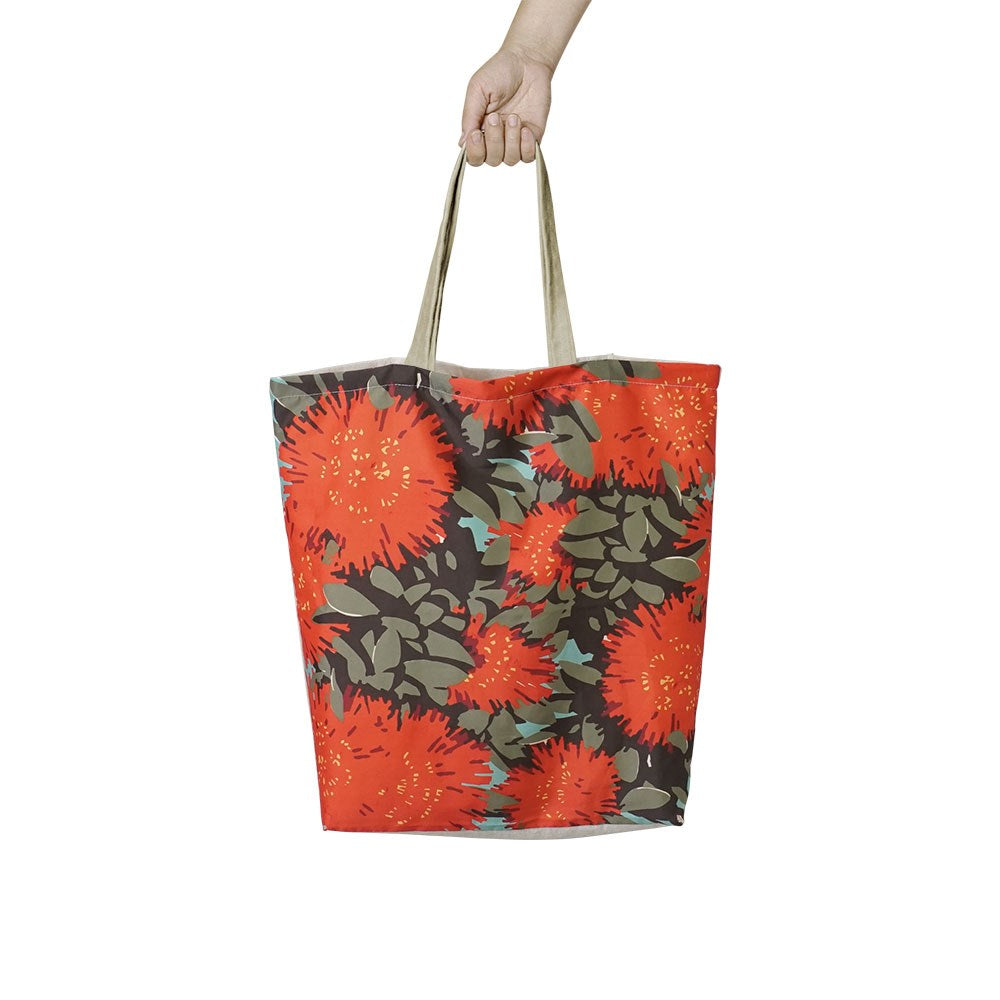 A hand holds the Vintage Pohutukawa Shopping Bag, a lightweight, affordable tote with tan handles and a bold red Pohutukawa floral pattern on a dark background—ideal as a reusable shopping bag.