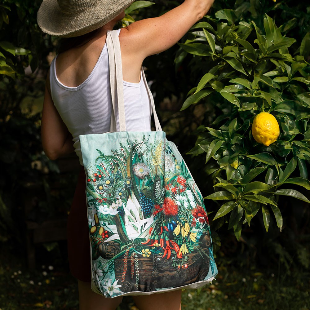 A person in a sunhat, white tank top, and red shorts stands outdoors by a tree with a yellow fruit, holding the affordable NZ Native Flowers Shopping Bag decorated with colorful floral artwork.