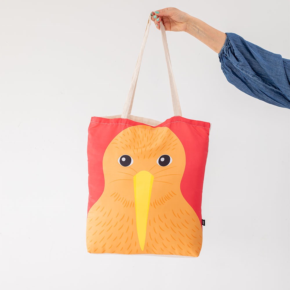 A person in a blue long-sleeve top holds the Bright Kiwi Shopping Bag, a lightweight tote with beige handles featuring an orange kiwi bird with a yellow beak on a red background.