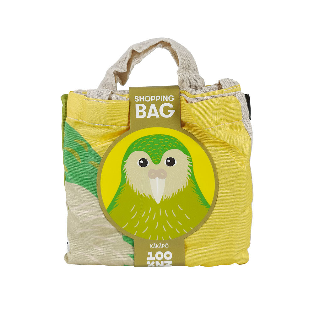 The Bright Kakapo Shopping Bag is an affordable, lightweight yellow and green reusable bag with short fabric handles and a circular cardboard tag featuring a green kakapo illustration. The tag reads SHOPPING BAG KĀKĀPŌ.