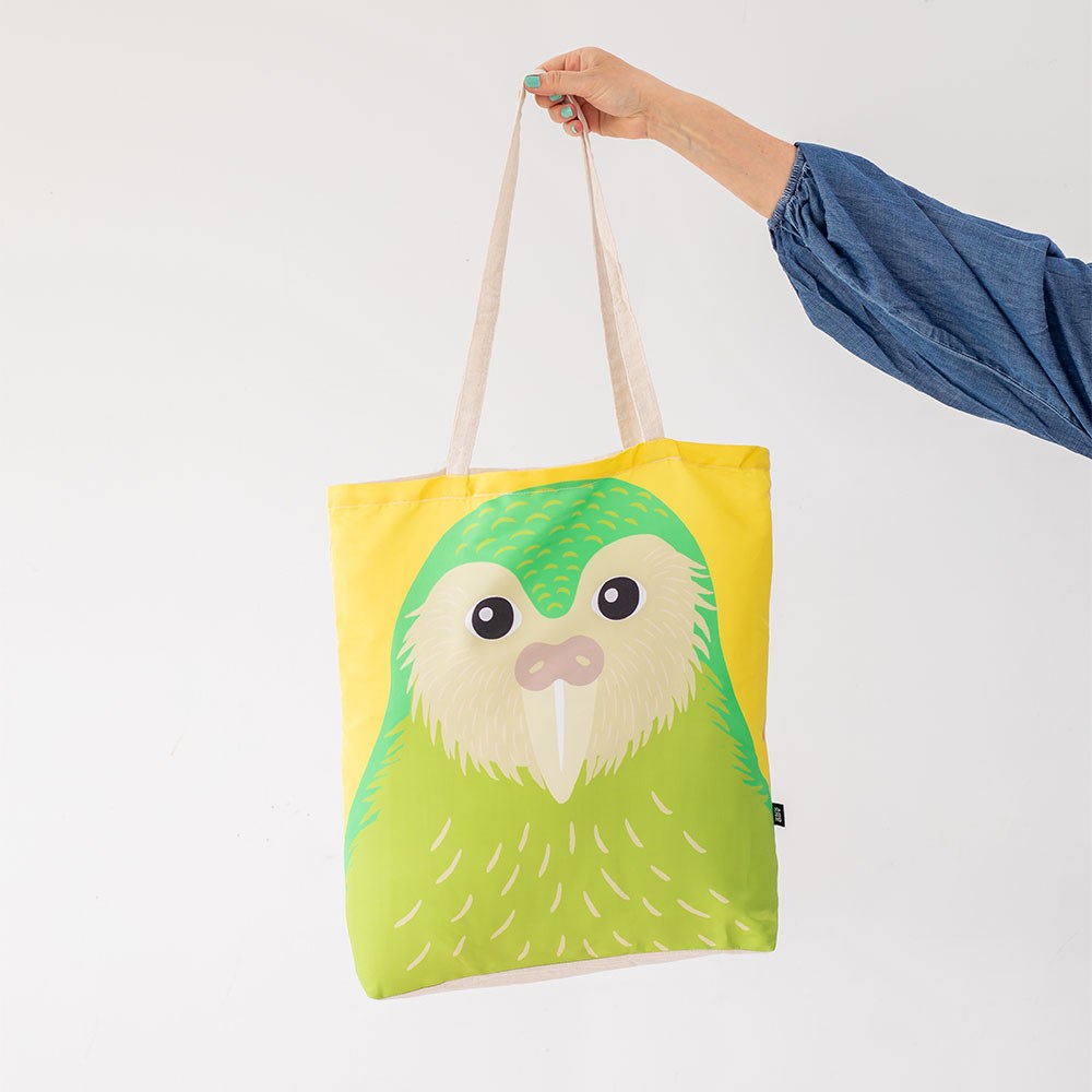 A hand holds the Bright Kakapo Shopping Bag with beige handles, showing a large green kakapo on a yellow background. The person’s blue sleeve is visible.