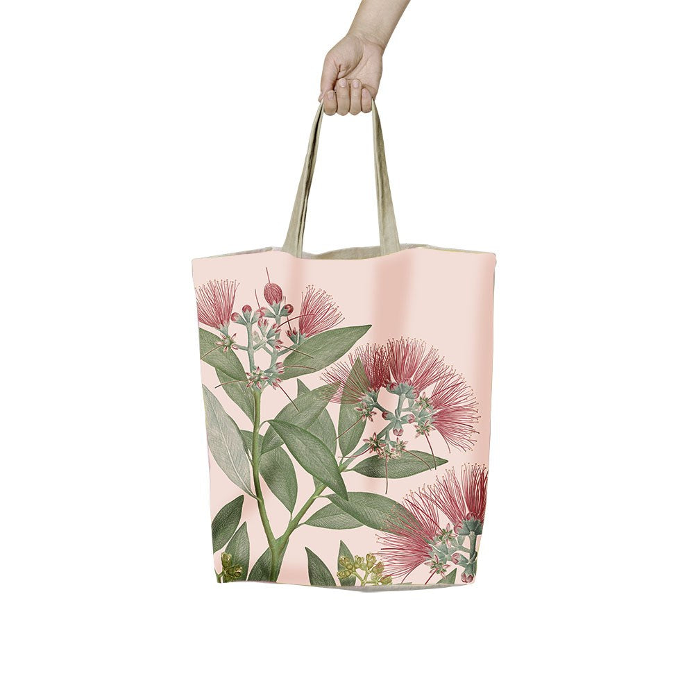 A hand holds the Vintage Botanical Pohutukawa Shopping Bag, an affordable beige tote with two handles and a green leaves and red flowers print on a light pink background—ideal for stylish, reusable shopping.