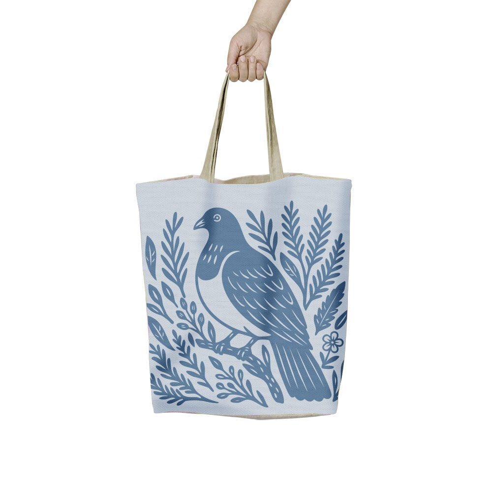 A hand holds a Woodcut Kereru Shopping Bag featuring a blue woodcut-style illustration of a kereru bird perched on a branch surrounded by foliage.