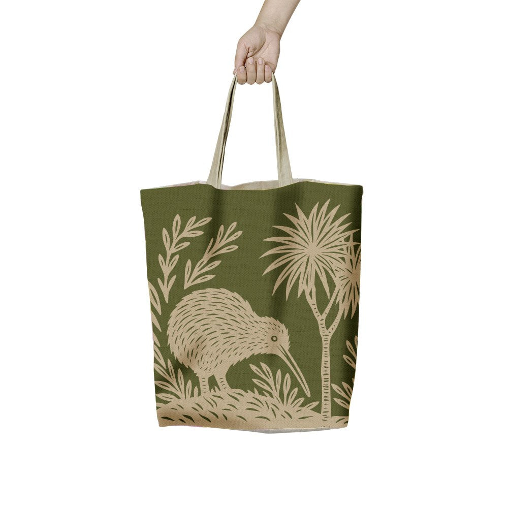 A hand holds a Woodcut Kiwi Shopping Bag featuring a beige woodcut-style illustration of a kiwi bird among tropical plants on a dark green background.