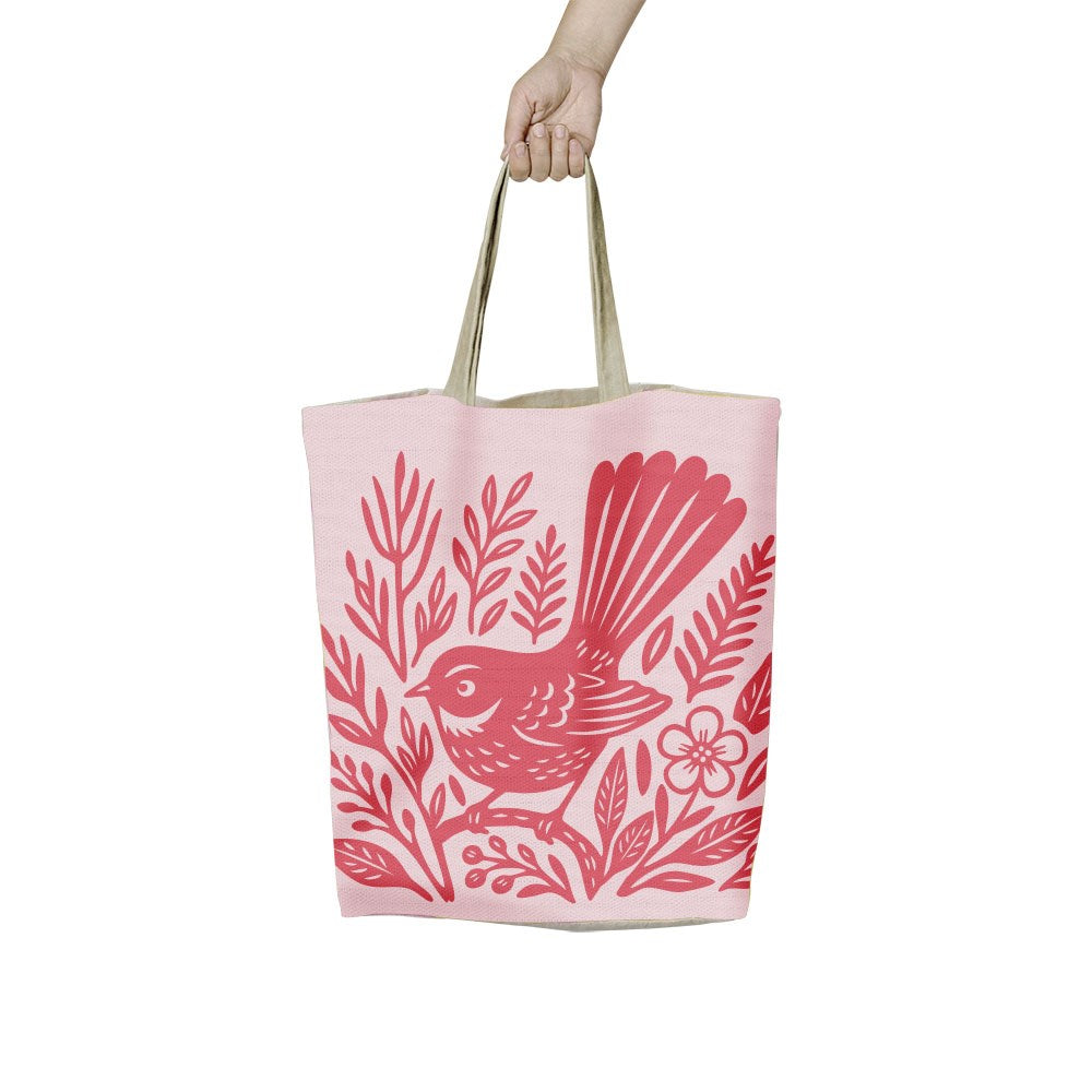 A hand holds a light pink Woodcut Fantail Shopping Bag featuring a red woodcut-style illustration of a fantail bird perched among flowers and leaves.