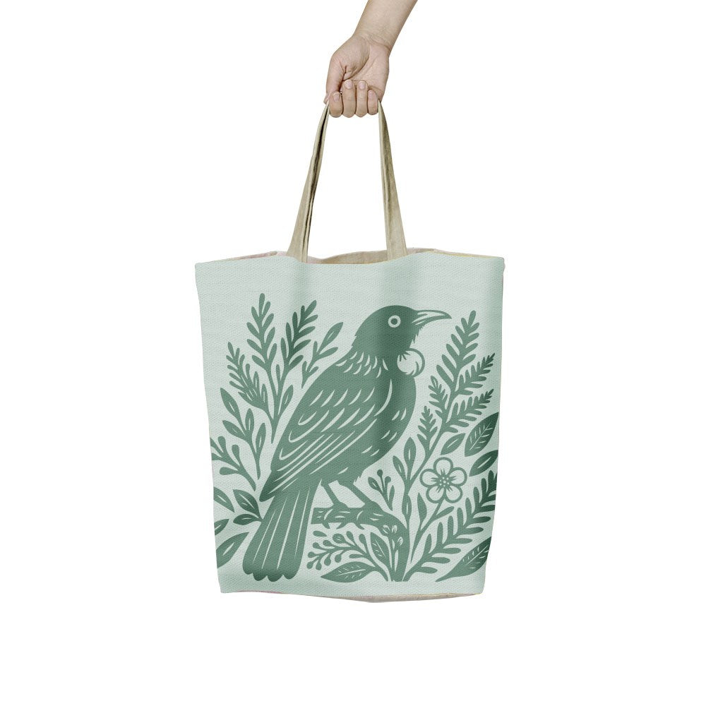 A hand holds a lightweight Woodcut Tui Shopping Bag, a New Zealand-designed canvas tote featuring a green woodcut-style illustration of a tui bird perched among foliage.