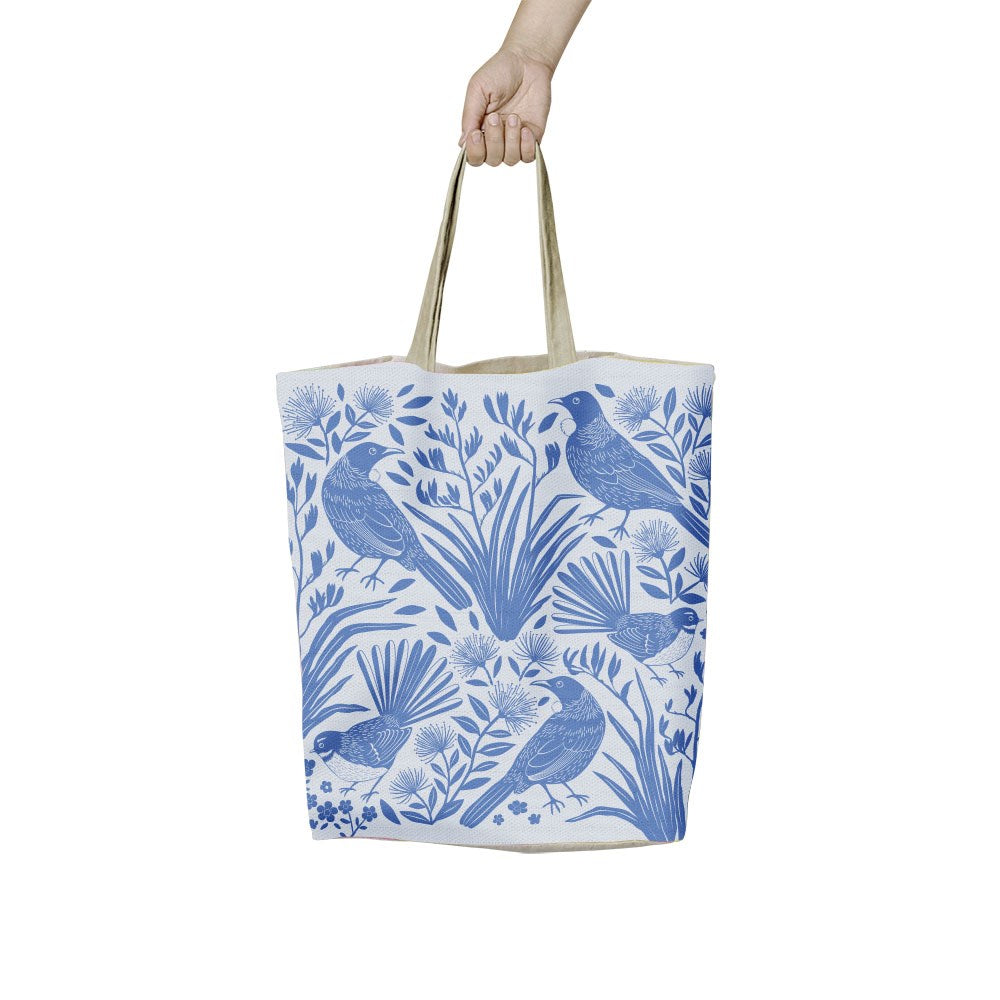 A hand holds a Birdsong Blues Shopping Bag featuring a blue and white pattern of birds and native plants.