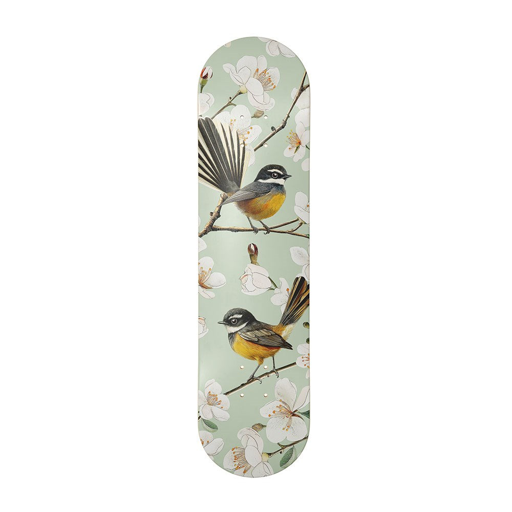 The Chatty Fantails Skateboard Deck features a contemporary illustration of two birds with yellow and brown bodies, accented by black and white, perched on flowering branches against a pale green background. Made from Canadian maple.