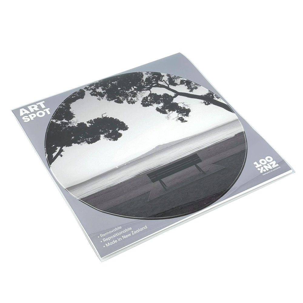 Square package with a round sticker featuring a black-and-white photo of Mission Bay bench by the water. Text reads Mission Bay Art Spot and highlights removable, repositionable peel-and-stick wall art. Made in New Zealand.